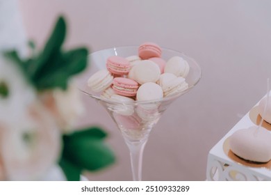 A glass bowl filled with pink and white macarons. The macarons are arranged in a way that they look like they are in a martini glass. The bowl is placed on a table next to a vase of flowers - Powered by Shutterstock