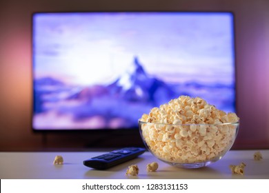 A glass bowl of popcorn and remote control in the background the TV works. Evening cozy watching a movie or TV series at home
