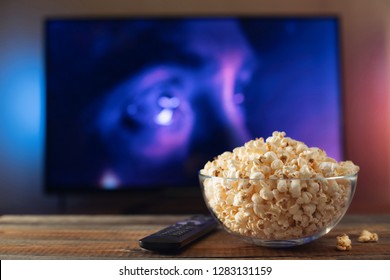 A glass bowl of popcorn and remote control in the background the TV works. Evening cozy watching a movie or TV series at home