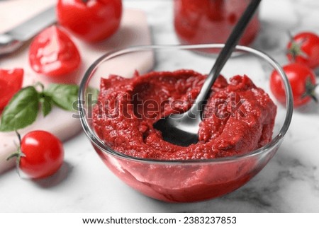 Glass bowl of tasty tomato paste with spoon and ingredients on white marble table, closeup