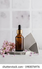 Glass Brown Serum Bottle With A Pipette For Skin Care With Branches And Flowers Of Lilac On A Gray Background. Health And Beauty Concept