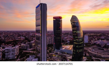 Glass Buildings In Europe City Life. Aerial View Of Modern Architecture Of Office Buildings In New Areas. Lights Of The Night City. The Drone Shoots At Three Towers. Sunset. Milan, Italy, August 2021: