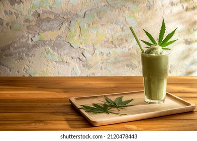 A glass of cannabis smoothie with cannabis leaves in a wooden tray on a wooden table over abstract colorful wall in warm light background. Copy space backdrop. Eatable vegetable healthy concept ideas.