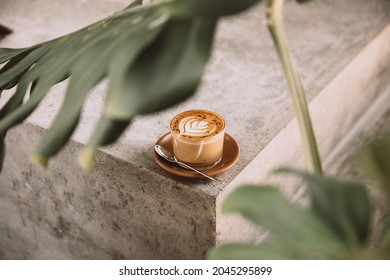 Glass of cappuccino with latte art on saucer and with spoon on the concreet background with tropical plant in front