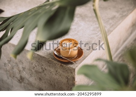 Glass of cappuccino with latte art on saucer and with spoon on the concreet background with tropical plant in front
