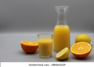 glass and carafe of illuminating yellow juice. cutted oranges and lemons nearby. ultimate gray background. natural drink with vitamin c. still life, copy space