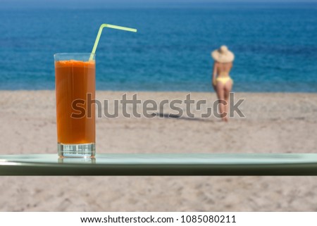 A glass of carrot juice with a straw in the cafe. Female in the bikini and straw hat on the beach 
