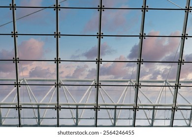 Glass ceiling panels and a supporting metal framework viewed from below, showing a partly cloudy sky with pink hues at sunset.
