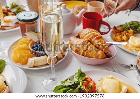 Glass of champagne close-up on the table with different breakfast and brunch dishes like croissants, eggs benedict, coffee and fruits. Selective focus