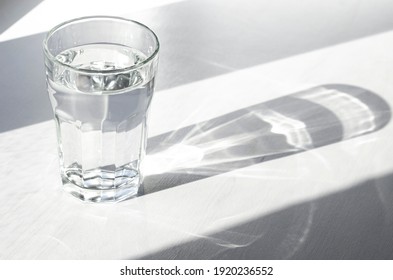 A glass with clean clear water and sharp shadows stands on a white wood table. top view of glass with water on white table