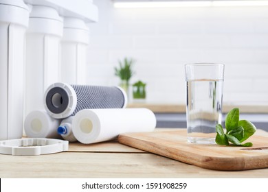 A glass of clean water with osmosis filter, green leaves and cartridges on wooden table in a kitchen interior. Concept Household filtration system.