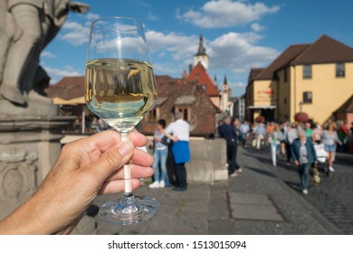 A Glass Of Clear White Wine Is Held Up Toasting The German City Of Wurzburg. Deliberately Blurred Background Of Tourists Walking And The Buildings. Inverted Reflection Of The City Skyline Can Be Seen 