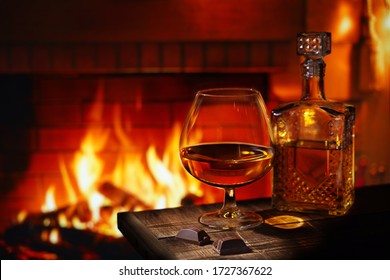 A glass of cognac with a bottle and chocolate on a wooden table near the burning fireplace. Concept winter evening, 
heat in the house, holiday. Free space for text.                               