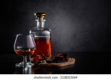 A glass of cognac, cognac in a bottle and pieces of chocolate on a dark background. Copy space.
