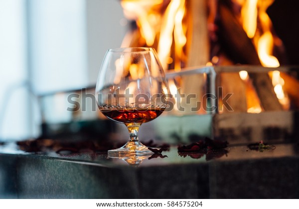 A glass of cognac on oak table on the\
background of a burning\
fireplace