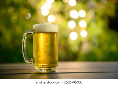 A Glass Of Cold Beer With A Foam On Top Placed On A Table In The Garden, The Background Is A Green Tree And Has Beautiful Bokeh At Dusk.