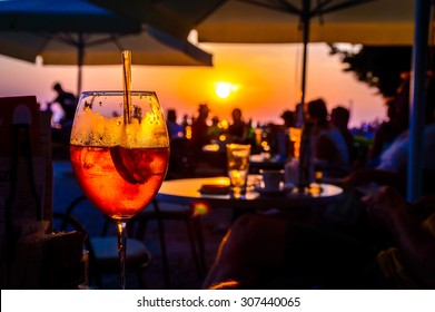 A glass of cold orange cocktail at the sunset on the table of a beach bar at the sunset, with blurry people around having refreshments or partying on a summer evening, with copy space for text