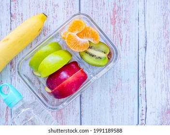 Glass Container Lunch Box Fresh Fruit With Apple, Kiwi, Banana, Orange And Water Bottle On Light Blue Wooden Background. Take Away Food. Top View With Copy Space, Healthy Eating Concepts.
