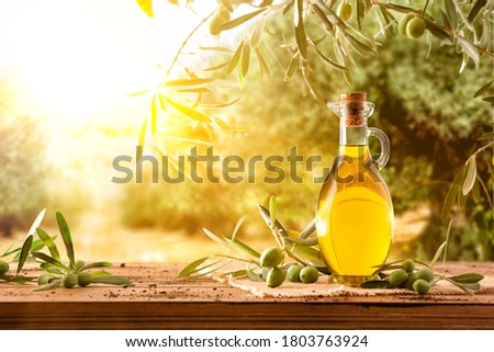 Glass container with olive oil on wooden table with branches and olives in crop field full of olive trees with sunshine