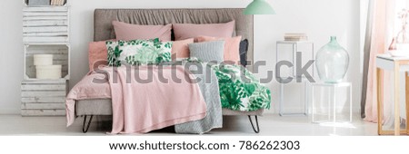Glass containers on white stools next to king-size bed with pink blanket in pastel cozy bedroom interior