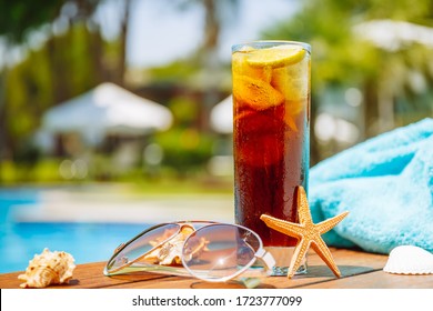 Glass Of Cuba Libre Cocktail With Seashells, Sunglasses And Blue Towel Near Swimming Pool. Vacation, Getaway, Holiday And Luxury Resort Concept. Close Up