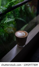 Glass Cup Of Coffee With Milk Shaped Leaf Sitting On A Wooden Ledge Near The Window.