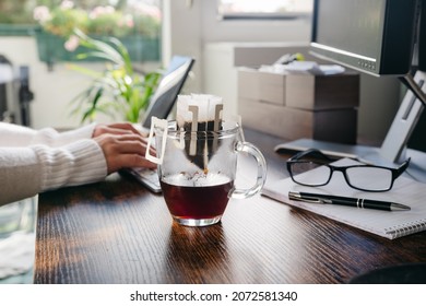 Glass Cup Of Freshly Brewed Coffee With Convenient Drip Coffee Bag On Dark Office Table. Womans Hands Typing On Keboard At The Background