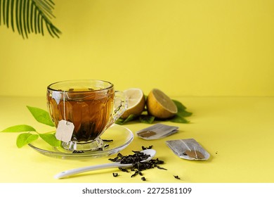 A glass cup with hot tea from a bag on a yellow table. Tea bags and lemon wedges in the background. Spoon with dried tea on the table. Light yellow background, text space - Powered by Shutterstock