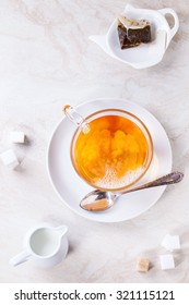 Glass Cup Of Hot Tea On Saucer With Sugar Cubes, Jug Of Milk And Tea Bag Over White Marble Backgtound. Top View