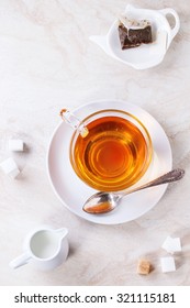 Glass Cup Of Hot Tea On Saucer With Sugar Cubes, Jug Of Milk And Tea Bag Over White Marble Backgtound. Top View