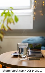 Glass Cup Of Hot Water On Wooden Table With Watercolor Drawing In Paper Notebook Near Home Flower And Bed In Bedroom Near Window And Lights On Cozy Cloudy Autumn Or Winter Day. Drink, Weekend, Holiday