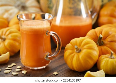 A glass cup of pumpkin juice. Pumpkins on kitchen table. Jug of pumpkin juice on background.