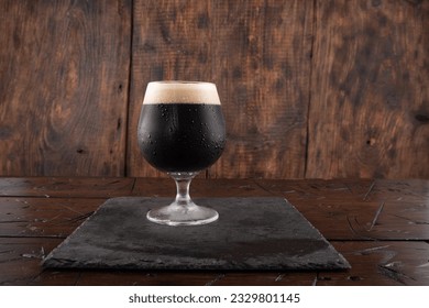 A glass of dark beer on a wood background. Beer on a slate tray. Serving stout beer. Front view.
