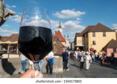 A Glass Of Dark Red Wine Is Held Up Toasting The City Of Wurzburg. Deliberately Blurred Background Of Tourists Walking And The City Buildings. Silhouette Of He City Skyline Can Be Seen In The Wine 