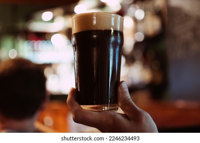 glass of dark stout beer with foam and bubbles in hand of a man in pub behind the bar