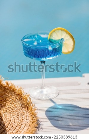 A glass of delicious blue cocktail Blue Lagoon and yellow lemons on the background of the pool. Alcoholic cocktail juicy fruit blue with curacao liqueur, ice cubes and a slice of lemon.