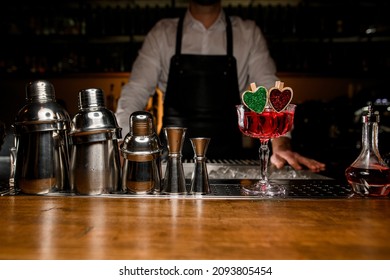 Glass with delicious tasty red alcohol cocktail decorated with small wooden green and red hearts standing on steel bar counter. Jiggers and shakers standing near.