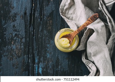 Glass of drawn ghee butter standing with wood spoon and linen textile over old dark blue wooden background. Healthy eating. Top view, copy space