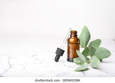 Glass dropper bottle with a pippette with black rubber tip and branch of eucalyptus near it on the beautiful white marble background. Nature Skin concept. Organic Spa Cosmetics. Trendy concept.