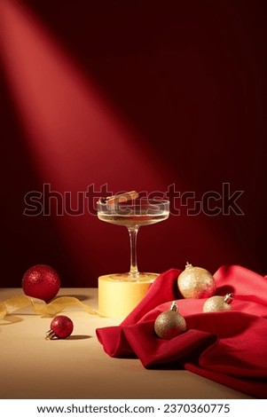 A glass filled with white wine and a cinnamon stick placed on yellow podium, displayed with baubles and red fabric. Another word for the Christmas season is Christmastime