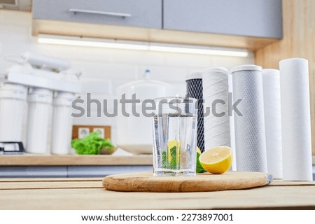 Glass of filtered clean water with reverse osmosis filter, lemons and cartridges on table in kitchen. Concept Household filtration or purification system.