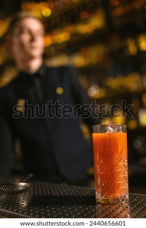 Glass with freshly made orange cocktail on counter against blurry barman view. Refreshing drink with ice cubes on metal table in pub