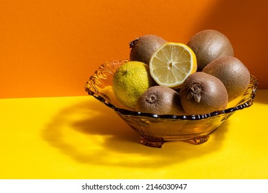 A Glass Fruit Bowl With Green Kiwi Fruits, Lime And A Sliced Lemon On A Yellow And Orange Background.