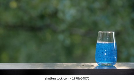 Glass full of with blue juice with jelly pieces on the brown organic wooden table with green morning summer or spring trees leaves bokeh background. Making drinks outdoors.