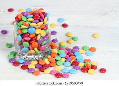 Glass full of colorful and sweet smarties. Chocolate buttons in colors of rainbow. Delicious candies on white wooden table.