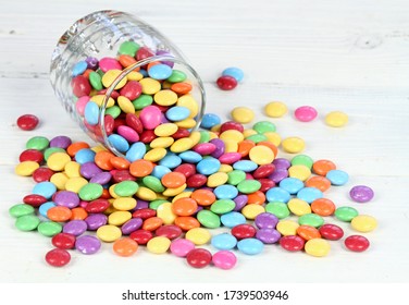 Glass full of colorful and sweet smarties.  Chocolate buttons in colors of rainbow.  Delicious candies on white wooden table.