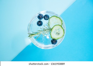 A Glass Of A Gin And Tonic Drink With Blueberries, Cucumber And A Rosemary Twig Isolated On Abstract, Colourful, Geometrical Color Block Blue Backgound