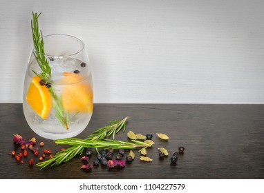 Glass Of Gin And Tonic With Ice, Orange And Rosemary Surrounded By Botanicals On Dark Wooden Surface