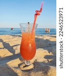 a glass goblet with a cool cocktail and a red tube stands in the sand on sea beach. quench your thirst while relaxing by the sea on a hot summer day
