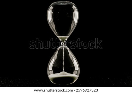A glass hourglass with sand in it is shown on a black background. The hourglass is almost empty, with only a small amount of sand left in it. Concept of time passing slowly